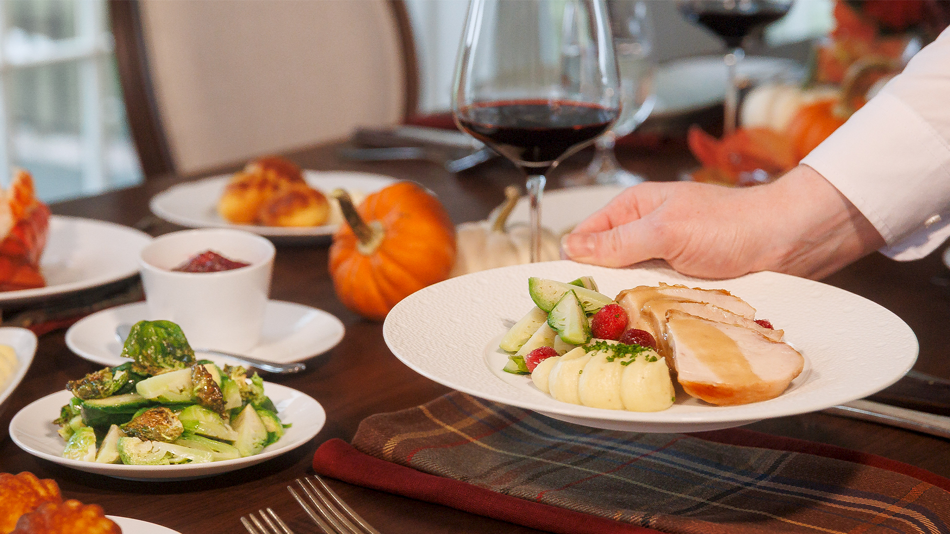 A chef sets down a plated meal: sliced meat with potatoes and greens, surrounded by other dishes at a table.