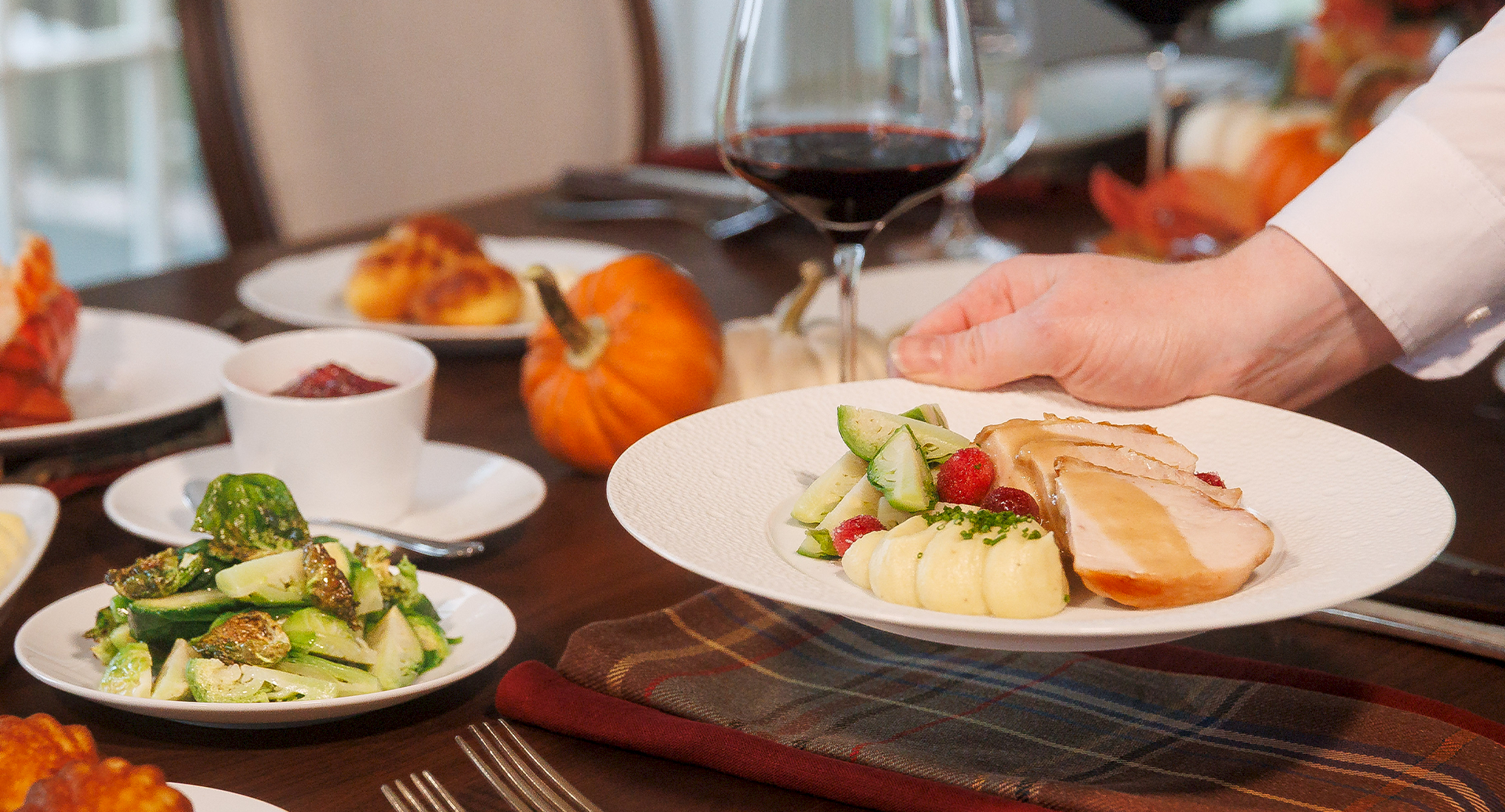 A chef sets down a plated meal: sliced meat with potatoes and greens, surrounded by other dishes at a table.