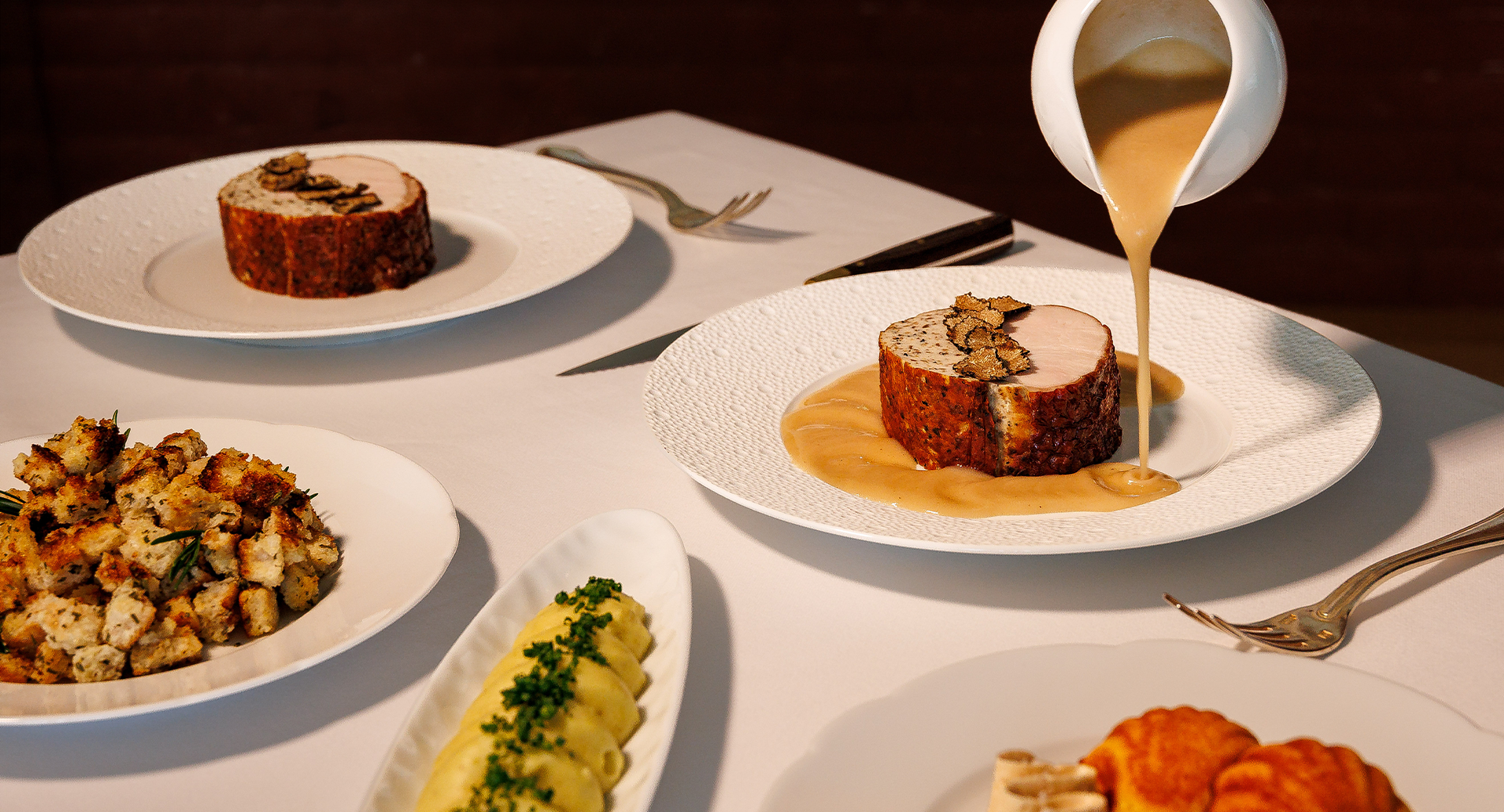 A fine-dining table with steak served on a plate as golden sauce is poured over it, alongside other gourmet dishes and cutlery.