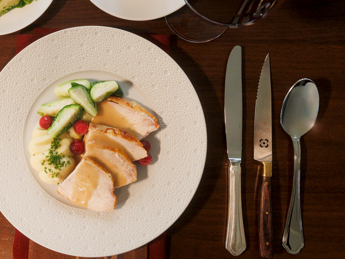 A plated meal with sliced chicken over creamy sauce, topped with cucumber and herbs, alongside veggies and rolls on the table.