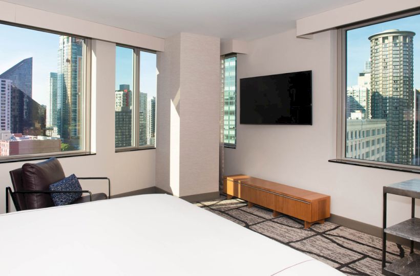A modern hotel room with large windows showcasing a cityscape, a wall-mounted TV, chair, and minimalist decor completes the scene.