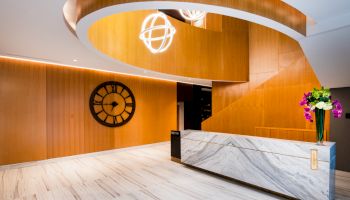The image shows a modern lobby with marble floors, a reception desk, large wooden staircase, wall clock, and decorative lighting.