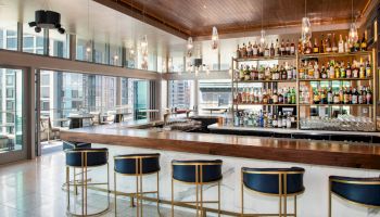 A modern, stylish bar with sleek stools, glass decor, and a well-stocked shelf of spirits, set against a city view.