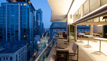A modern balcony overlooking a cityscape at dusk, with tables and chairs, and tall buildings visible along a street.