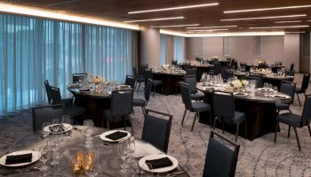 The image shows an elegantly set banquet hall with round tables, chairs, glassware, and floral centerpieces, under soft lighting.