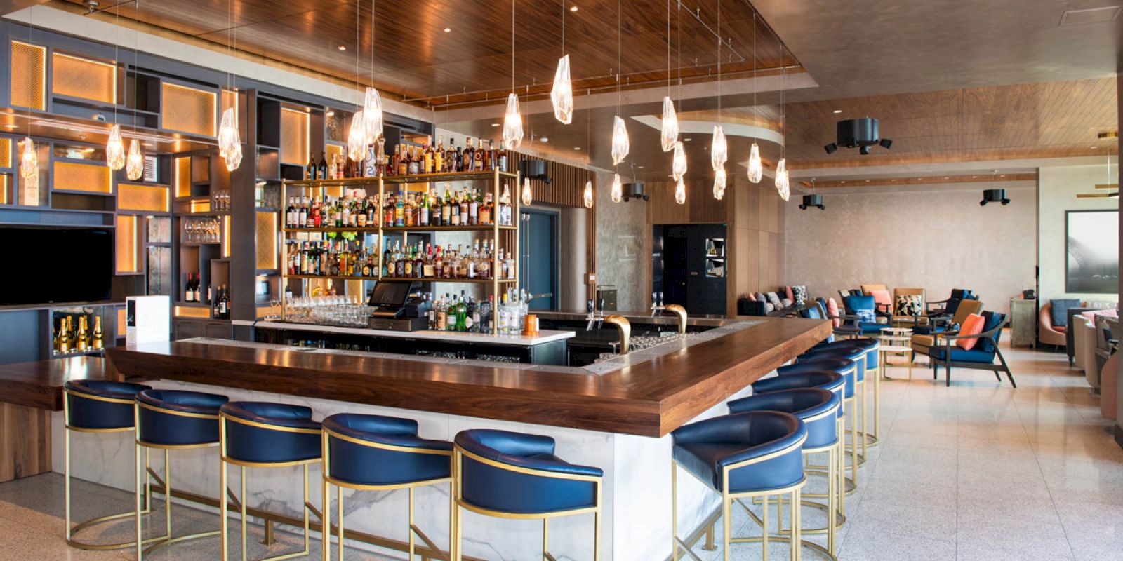 A modern bar with a stocked shelf, pendant lights, and blue bar stools. The seating area is in the background with a stylish design.