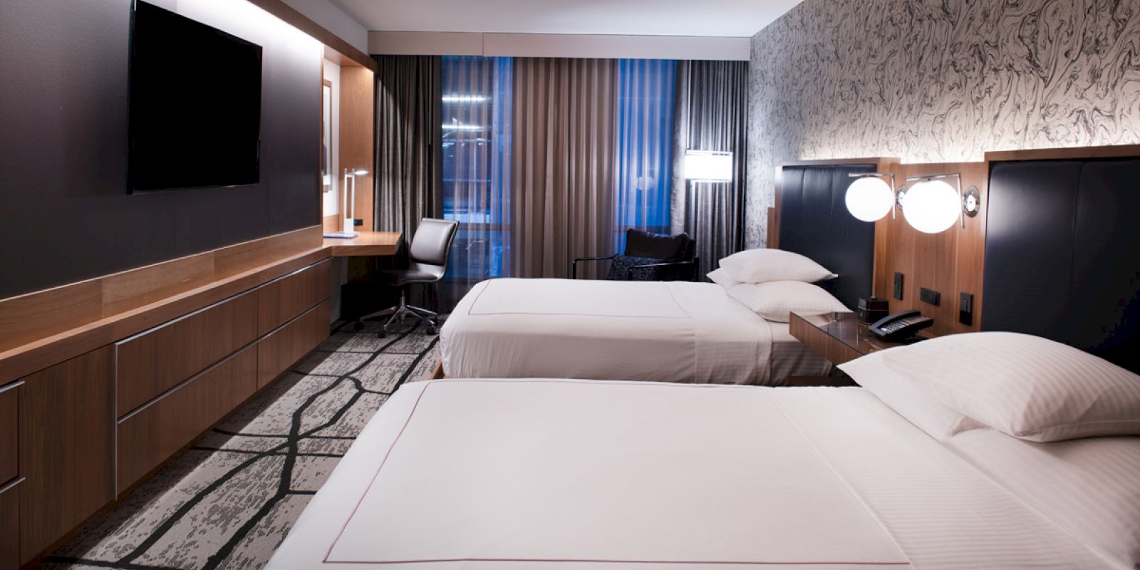 The image shows a modern hotel room with two double beds, a flat-screen TV, a work desk, lamps, and a patterned wallpaper.