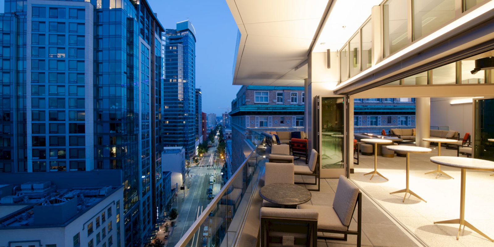 The image shows a modern rooftop terrace with seating, overlooking a cityscape of tall buildings at dusk.