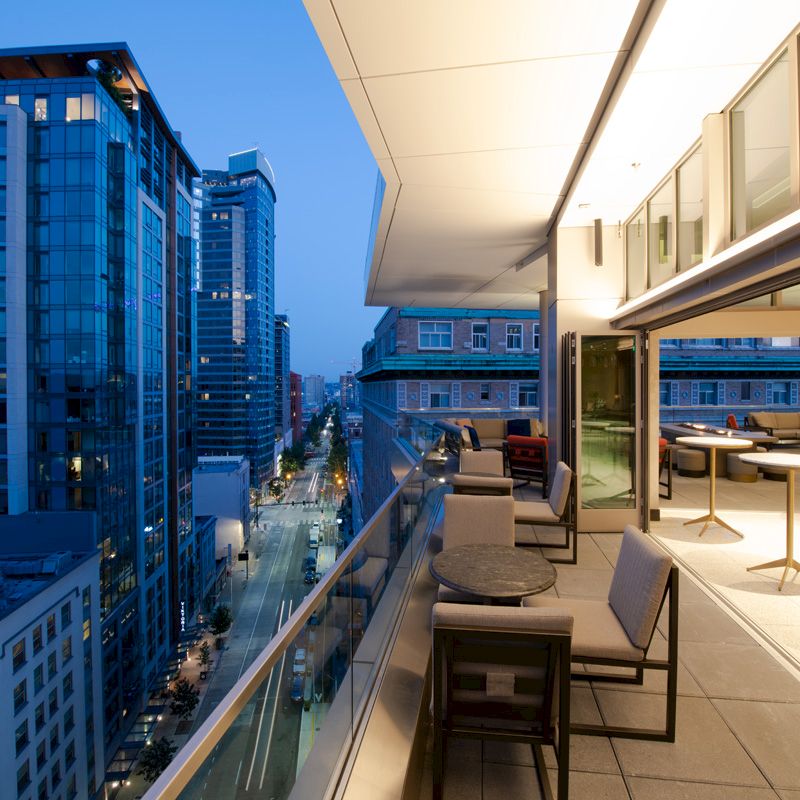 A modern cityscape at dusk is shown, featuring a balcony with seating, overlooking a street lined with tall buildings.