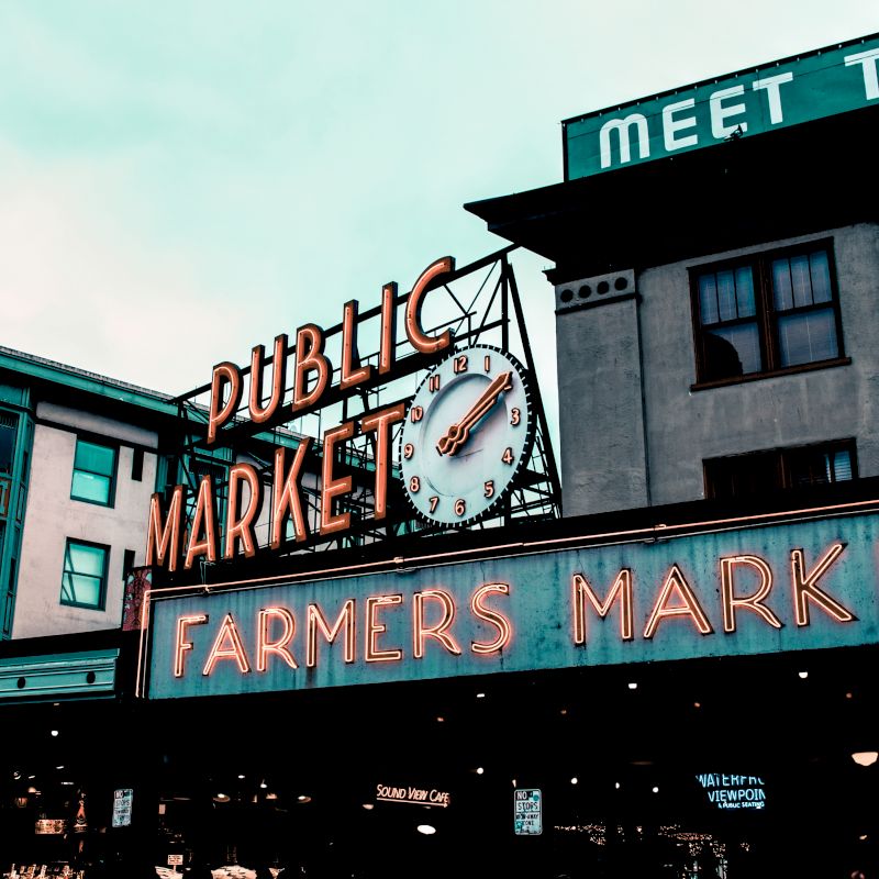 The image shows the entrance to a public market with a neon sign reading "FARMERS MARKET" and a clock above it, set against urban buildings.