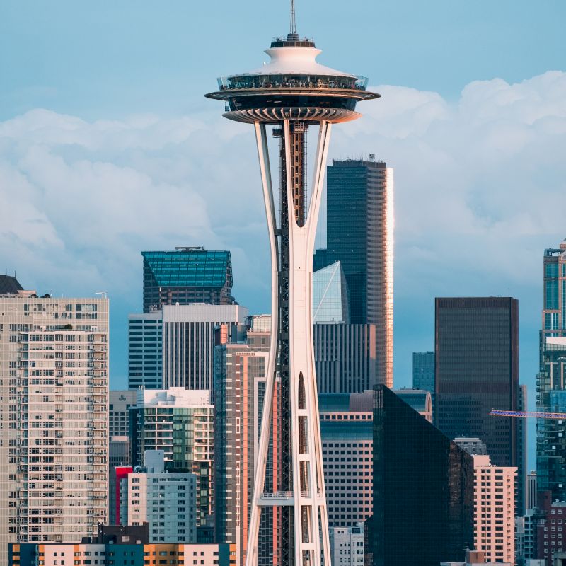 The image shows an urban skyline with the Space Needle prominently featured among various modern buildings and a cloudy sky backdrop.