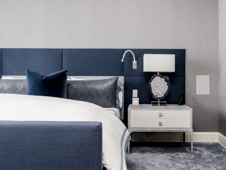 A modern bedroom with a blue bed, white bedding, navy pillows, and a bedside table with a lamp against a gray wall.