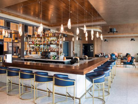 The image shows a modern bar with a wooden counter, blue stools, hanging lights, and shelves filled with bottles. There's seating in the background.