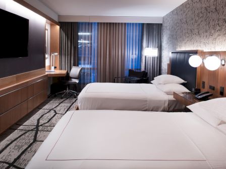The image shows a modern hotel room with two beds, a desk, a TV, and contemporary decor including patterned wallpaper and carpet.