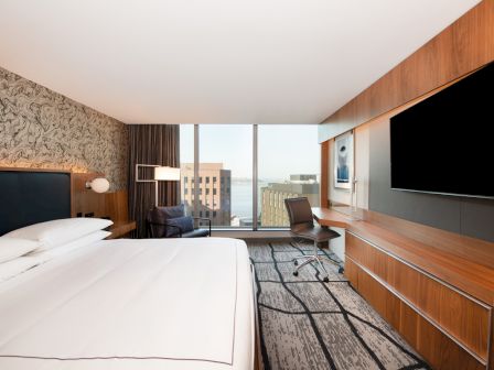 A modern hotel room with a bed, desk, and large TV, featuring a floor-to-ceiling window with an urban view.