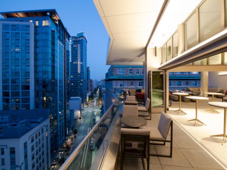 A modern cityscape with high-rise buildings at dusk, featuring a balcony with seating and tables overlooking the street below.