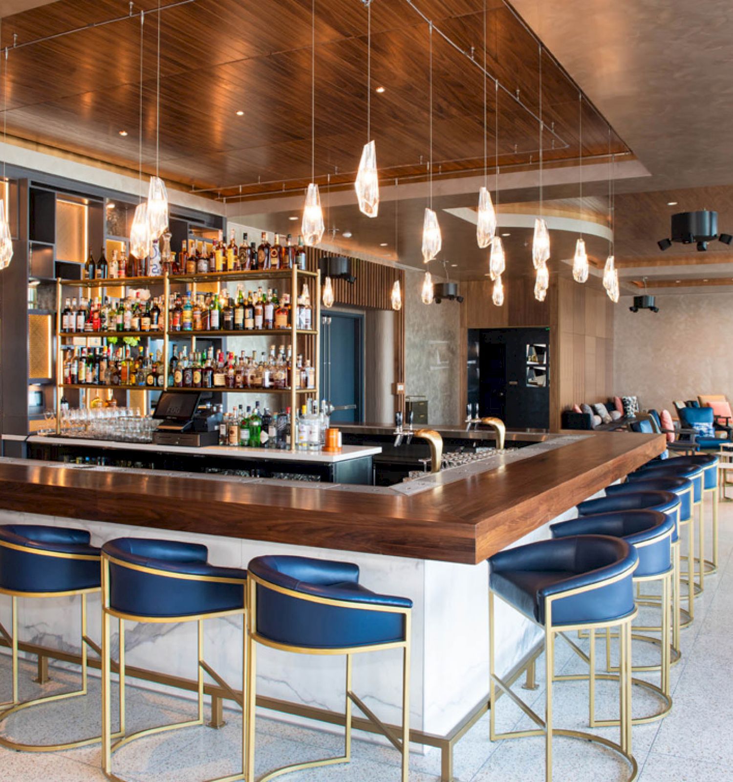 A modern bar with blue stools, hanging lights, and a well-stocked shelf. Cozy seating area in the background completes the scene.