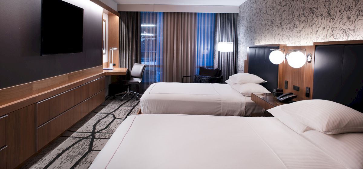 The image shows a modern hotel room with two beds, a wall-mounted TV, a desk, and contemporary decor, featuring a patterned accent wall.