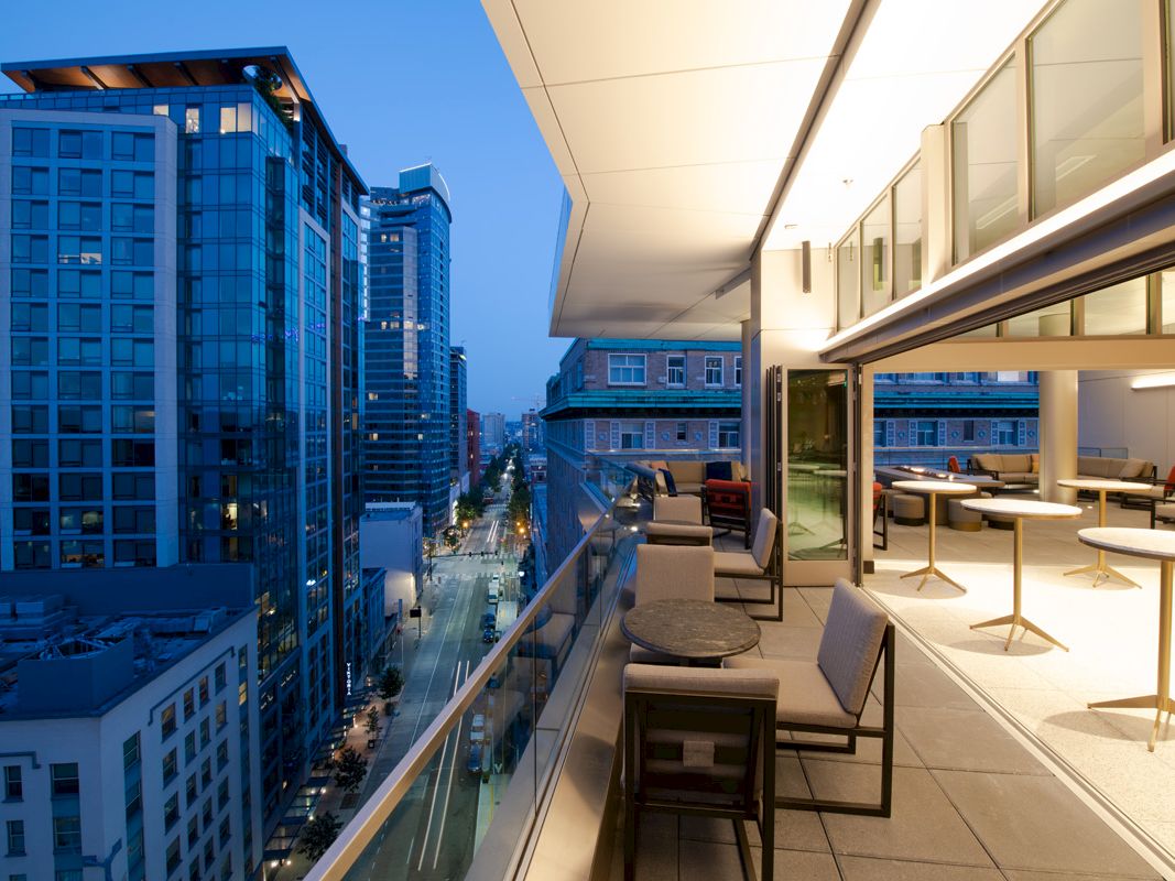 A modern cityscape with tall buildings and a well-lit balcony featuring tables and seating at dusk.