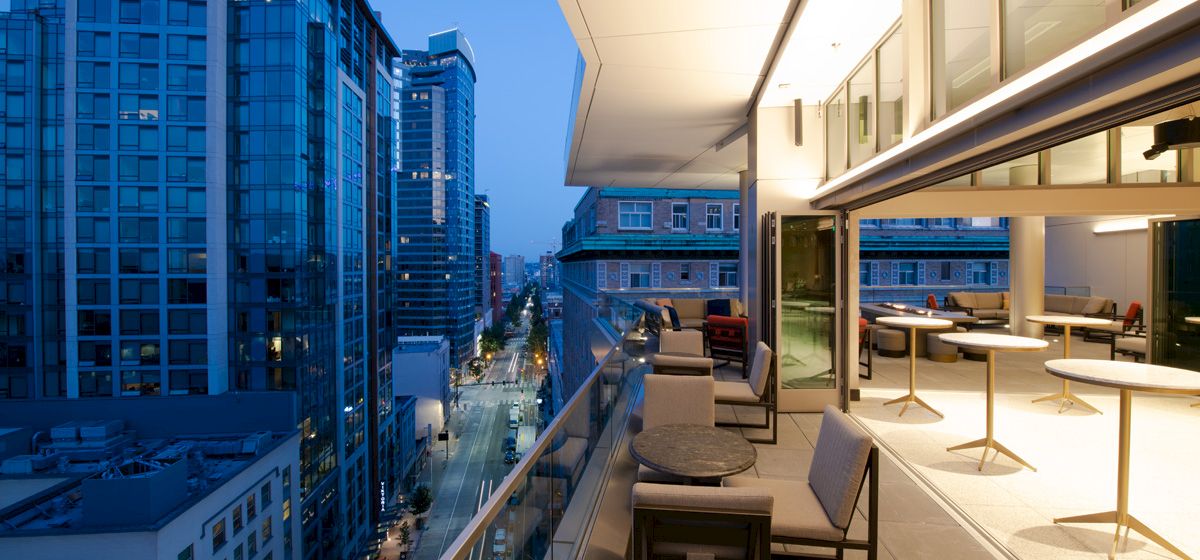 A modern cityscape with tall buildings and a well-lit balcony featuring tables and seating at dusk.