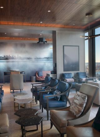 A modern lounge with comfy chairs, a skyline mural, large windows offering a waterfront view, and stylish lighting, creating a relaxing vibe.