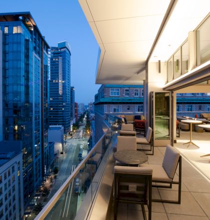 A modern balcony view at dusk shows city buildings with lit windows and a sleek outdoor dining area with tables and chairs.