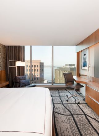 A modern hotel room with a large bed, wall art, desk, chair, wide window with city view, and a flat-screen TV on a wooden wall unit.
