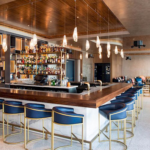 A modern bar with a sleek counter, blue stools, pendant lights, and a well-stocked shelf of bottles in a stylish lounge setting.