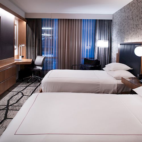 The image shows a modern hotel room with two beds, a TV, a desk, and a chair, featuring elegant lighting and decor.