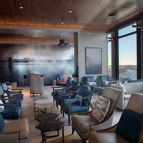 A modern lounge with plush seating, large windows showing a waterfront view, and a city skyline mural on the wall.