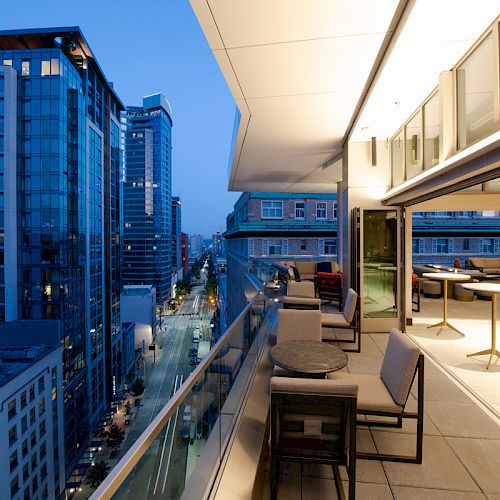 A modern cityscape at twilight shows a balcony view with tables and chairs overlooking high-rise buildings and a street below.