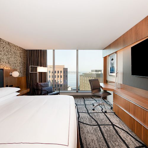 A modern hotel room with a large bed, desk, TV, and city view through a window. Contemporary decor with wood accents and patterned wallpaper.