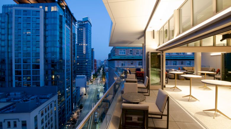 A modern cityscape at dusk with high-rise buildings, and an open balcony featuring tables and chairs along the street.