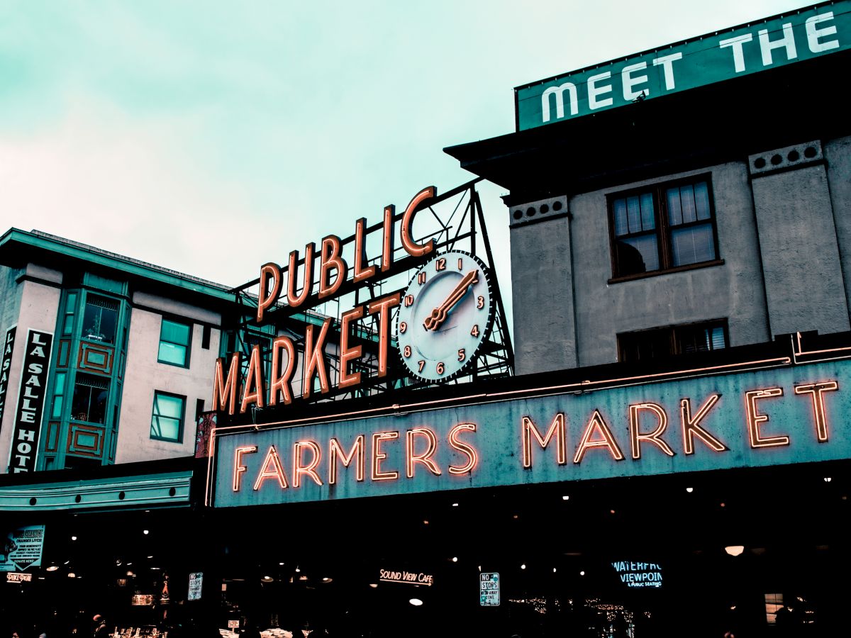 The image shows a neon sign reading "Public Market" and "Farmers Market" against a background of city buildings and a cloudy sky.