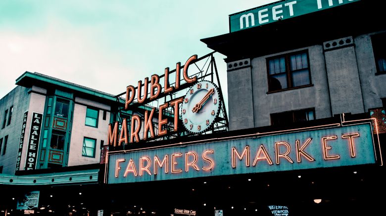 The image shows a neon sign reading "Public Market" and "Farmers Market" against a background of city buildings and a cloudy sky.