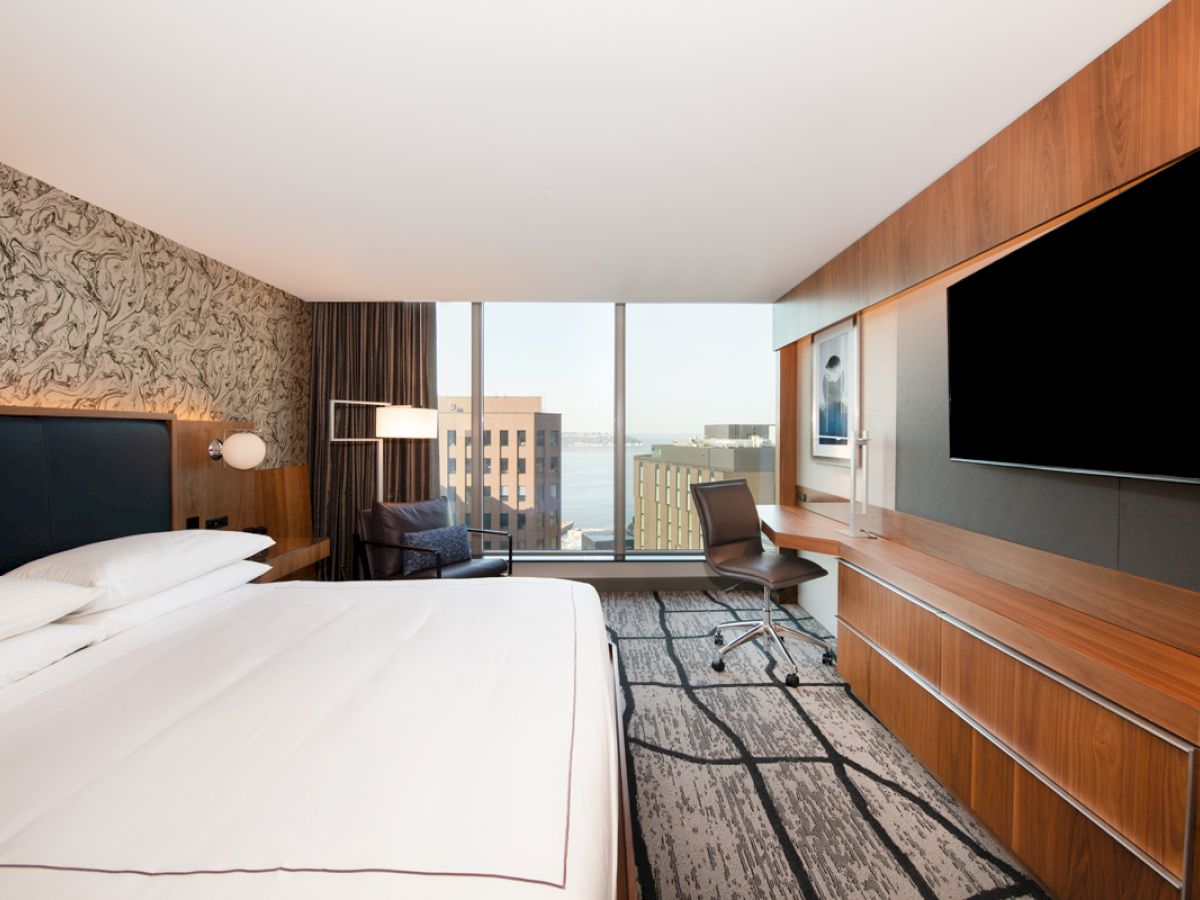 A modern hotel room with a bed, desk, chair, large TV, and a window with a city view.