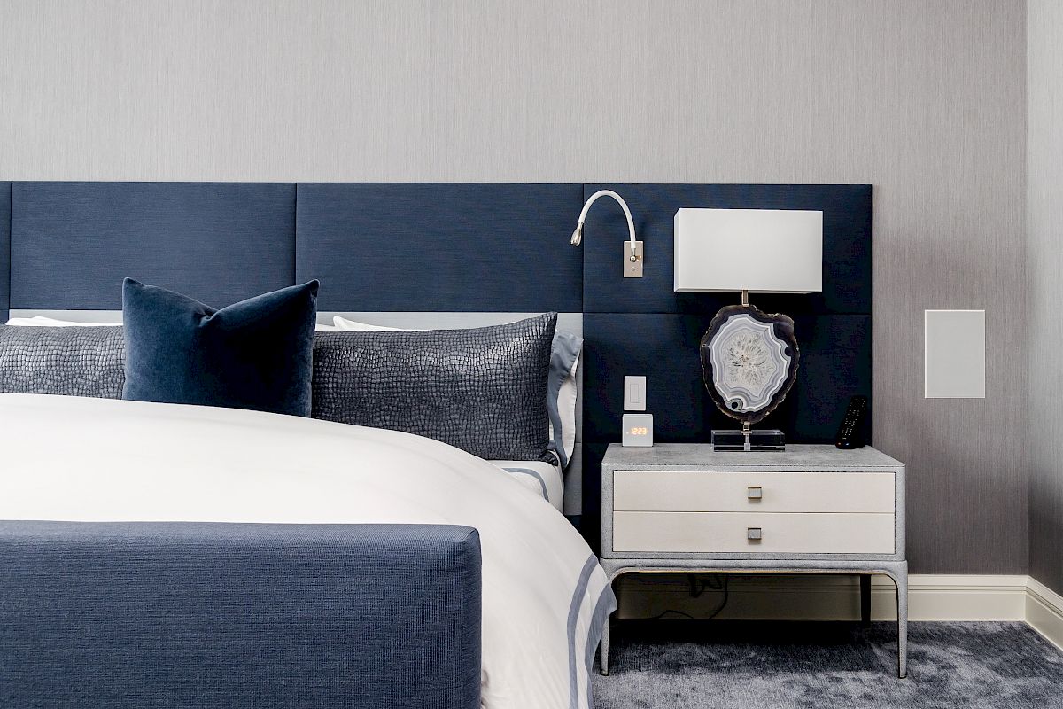 A modern bedroom features a blue upholstered bed, a grey and white nightstand with a lamp, and a plush carpet, creating a cozy atmosphere.