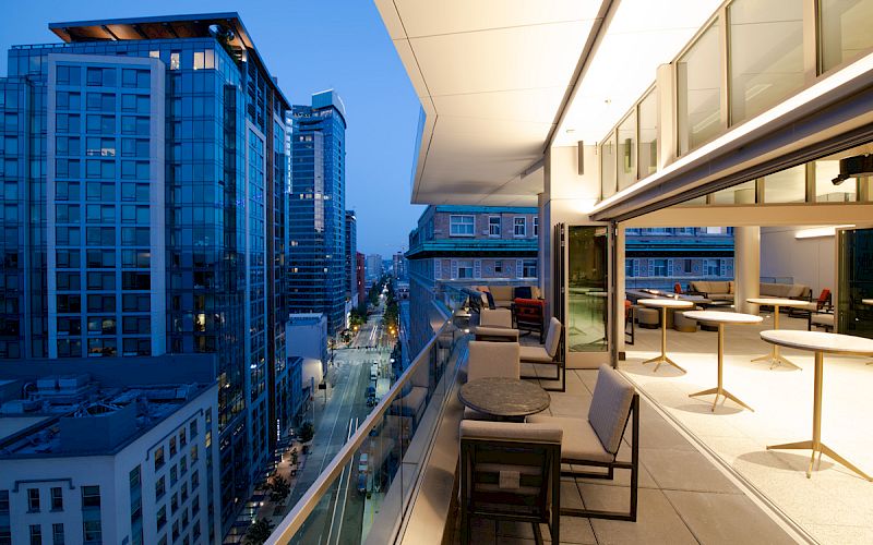 A modern rooftop terrace overlooking a cityscape at dusk, featuring outdoor seating and adjacent tall buildings.
