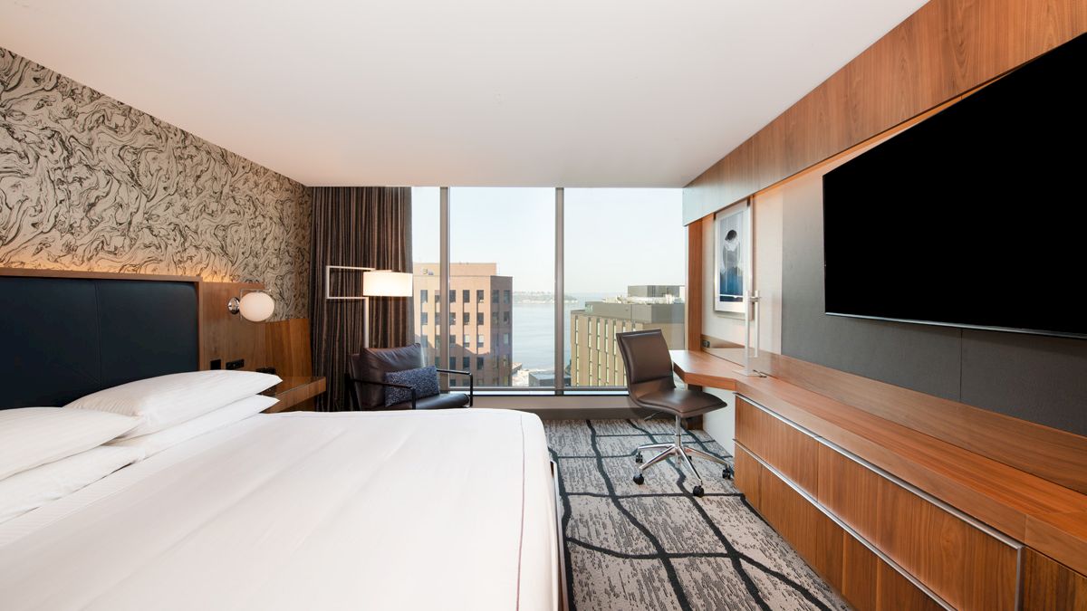A modern hotel room with a large bed, TV, desk, chair, and a window view of buildings and water outside.