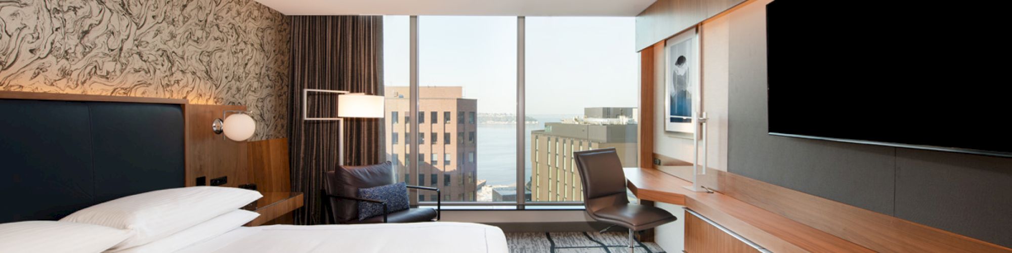 A modern hotel room with a large bed, TV, desk, chair, and a window view of buildings and water outside.