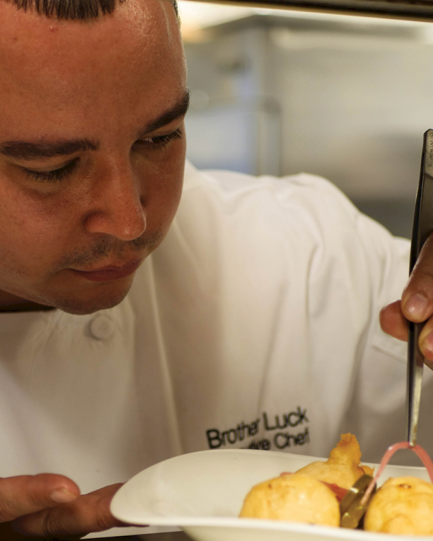 A chef in a white jacket tastes food with a spoon, concentrating over a plated dish in a restaurant kitchen.