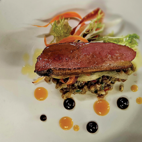 A plated steak medallion on greens and vegetables with herb garnish and colorful sauce dots around it.