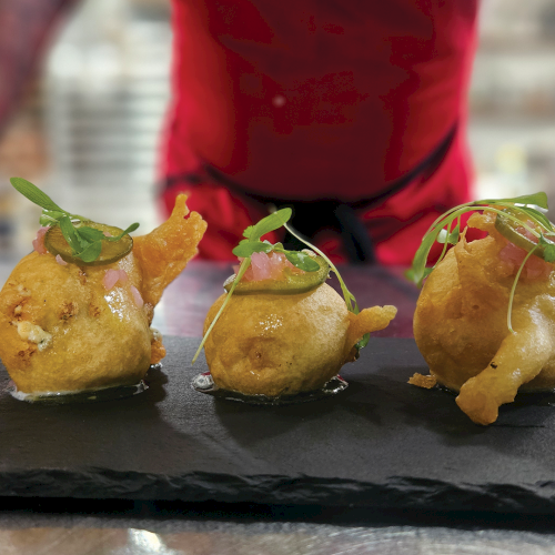 Three golden fried dumplings sit on a dark slate, garnished with herbs, while a chef in red prepares behind them.