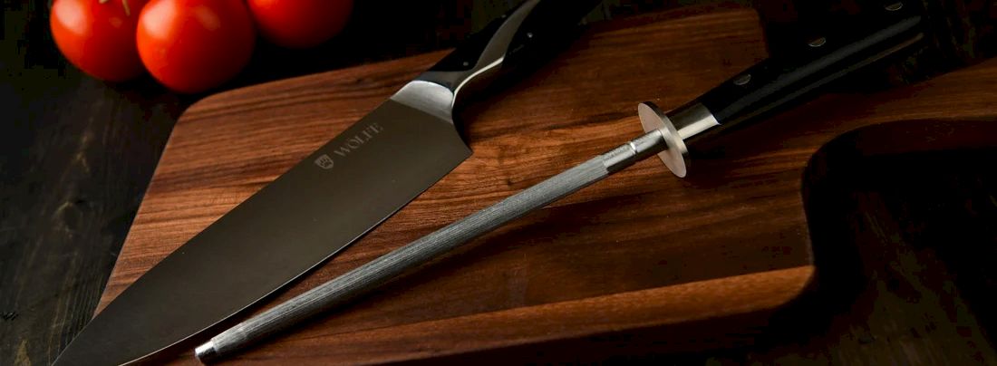 A chef&rsquo;s knife on a wooden cutting board beside red tomatoes on a dark countertop.