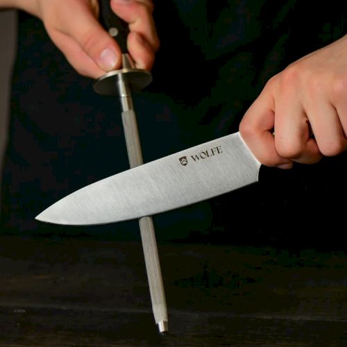 A person grips a WOLFE kitchen knife by its handle, blade angled over a dark table.