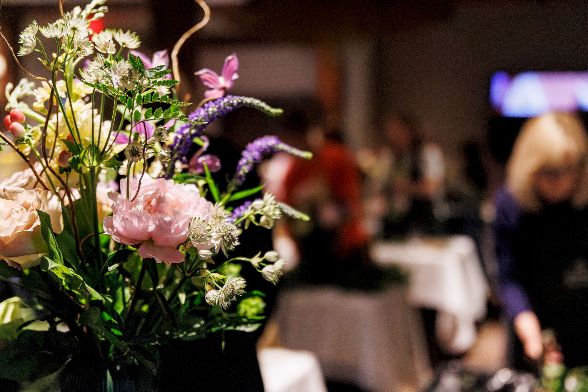 A bouquet of colorful flowers fills the foreground while a person is blurred in the background, holding or arranging the blooms.